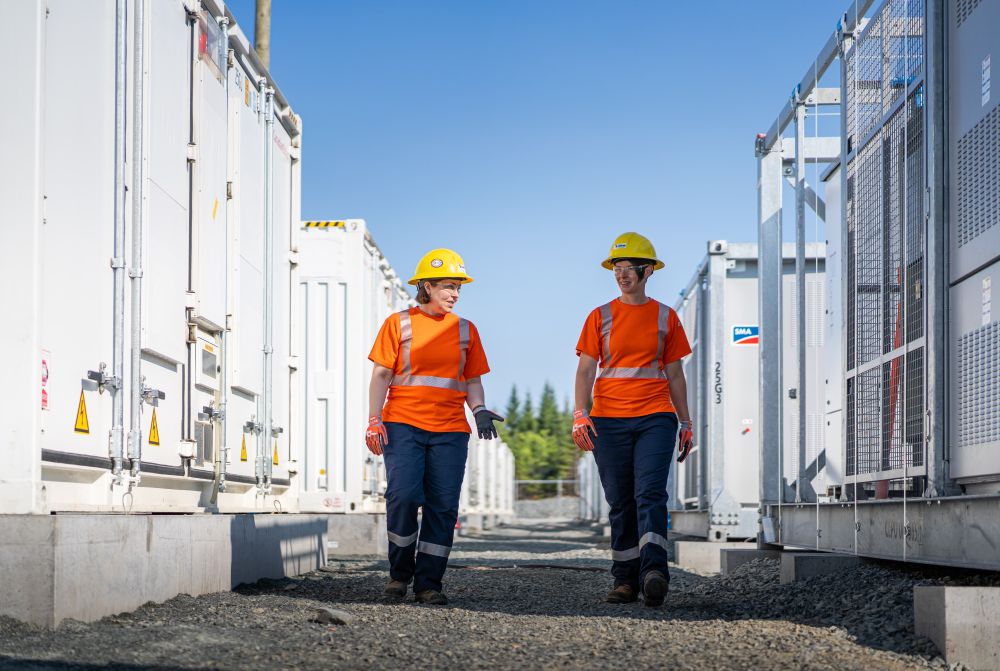 Two NS Power workers at the Waverley grid scale battery site wearing protective orange clothing, helmets, and golves